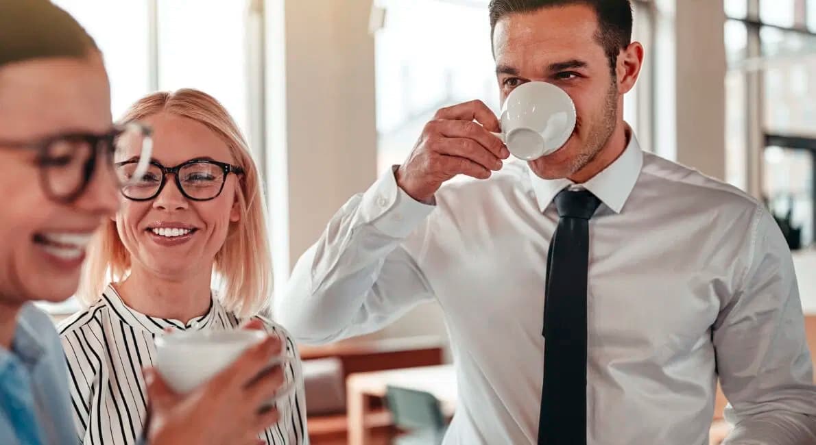 Tre personer i forretningstøj står sammen, smiler og holder kaffekopper i et lyst, moderne kontormiljø. En person drikker af en kop, mens de andre griner og sludrer.
