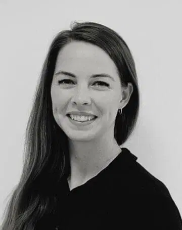 En kvinde med langt, glat hår smiler til kameraet. Hun er iført en mørk top og små øreringe. Baggrunden er ensfarvet og lys. Billedet er i sort og hvid.