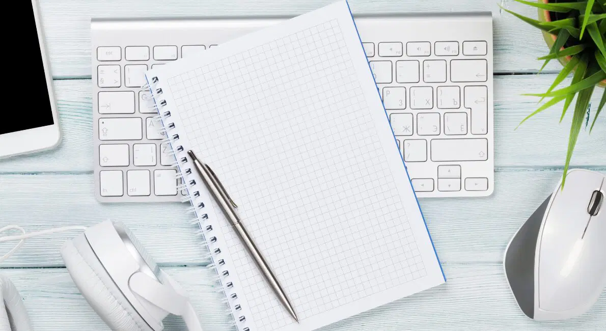 En tom notesbog med grafpapir og en pen hviler på et hvidt computertastatur. I nærheden ligger hvide hovedtelefoner, en trådløs mus, en grøn plante og hjørnet af en tablet, alt sammen på et lyst træbord.