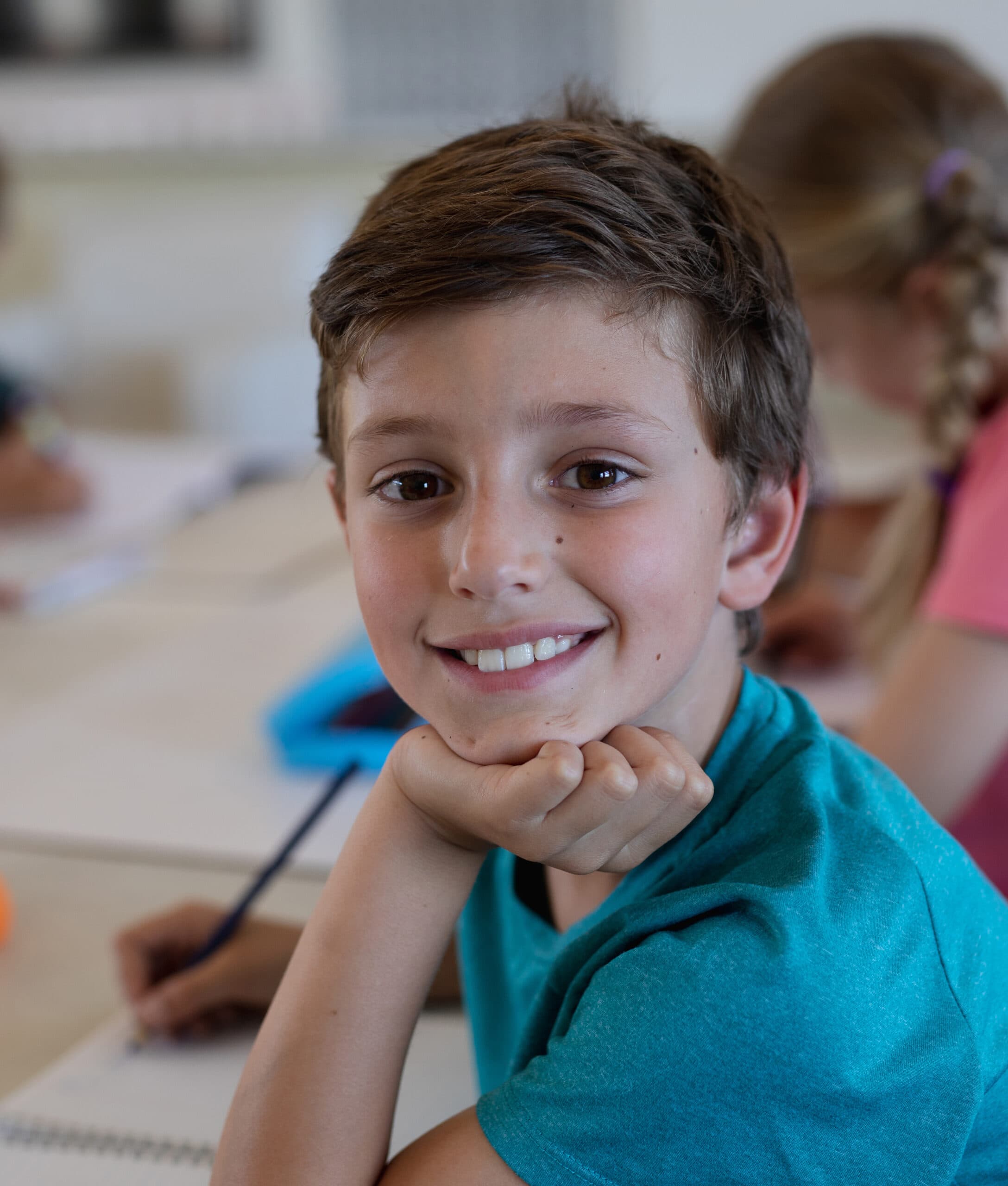 En smilende dreng med kort, brunt hår og en blågrøn skjorte sidder ved et skrivebord og hviler hagen på hånden, mens han skriver i en notesbog. Andre børn er slørede i baggrunden.